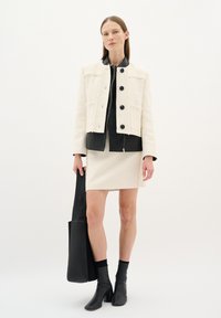 Giacca di tessuto crema con colletto in pelle nera e quattro bottoni neri. Abbinata a una camicia nera con zip, gonna crema e una borsa a tracolla nera.