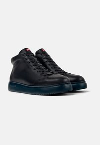 Zapatillas altas de cuero negro con un acabado suave, que presentan una puntera redonda, parte delantera con cordones y una suela oscura. Etiqueta de logo roja en la parte superior.