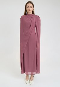 Długa, mauve sukienka z drapowanym materiałem, wysokim dekoltem i dopasowaną sylwetką. Posiada boczne rozcięcie i przezroczysty overlay, połączona z białymi szpilkami.