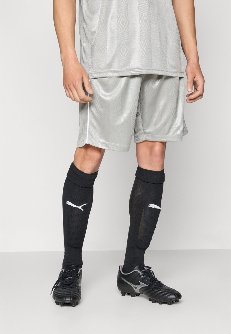 Camisola atlética cinza com padrão geométrico, calções cinzentos a condizer, meias pretas até ao joelho com logótipo e sapatilhas de futebol pretas com acabamentos em prata.