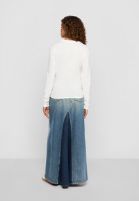 Vrouw met krullend haar staat met haar rug naar de camera, gekleed in een witte langemouwen top en een twee-kleurige denim maxi rok met een split aan de achterkant.