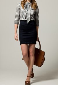 Donna che indossa una blusa a scacchi, una gonna nera e sandali con zeppa marroni, che porta una grande borsa marrone, in piedi davanti a uno sfondo semplice.