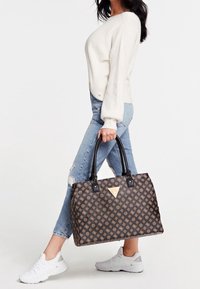 Femme portant un pull blanc et un jean bleu déchiré, marchant et tenant un grand sac fourre-tout Guess marron à motifs avec des poignées noires.