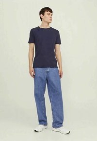 Man wearing navy t-shirt, loose blue jeans, and white sneakers standing against plain light background.