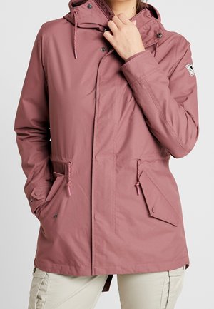 Vrouw draagt mauve kleurige capuchonjas met aantrekkoorden en klepzakken met knopen, houdt kraag met één hand vast.