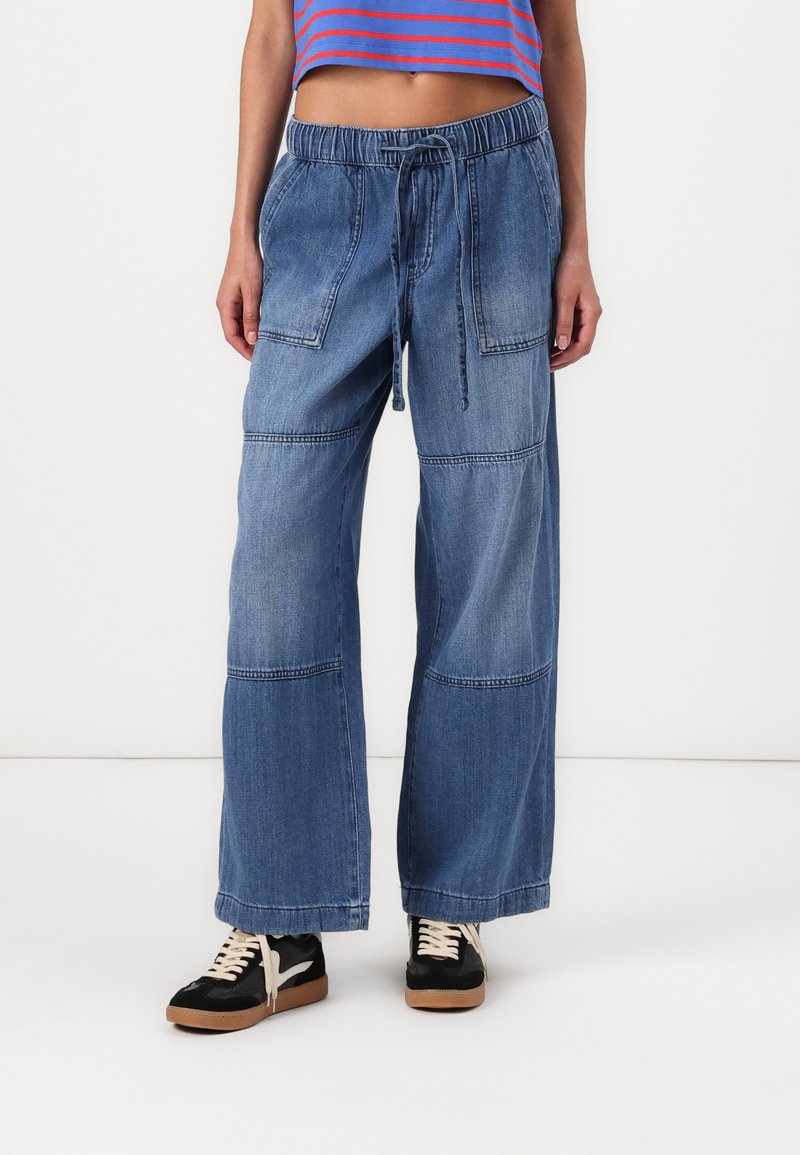 Lange blauwe denim broek met een drawstring tailleband, voorzien van patchzakken en contrasterende naden. Gecombineerd met zwarte sneakers.