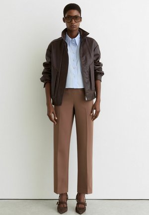 Oseba, oblečena v preveliko rjavo usnjeno jakno, svetlo modro srajco z gumbi, rjave hlače, rjave pete in velike temne sončna očala.