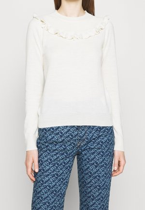 Žena v bílém svetru s dlouhým rukávem a volánky na hrudi a modrých vzorovaných kalhotách s opakujícím se nápisem.