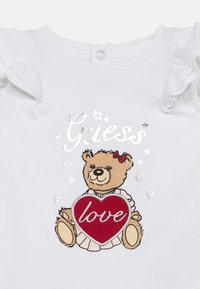 Body en coton blanc avec des manches à volants, arborant un motif d'ours tenant un cœur rouge marqué du mot "amour" en texte argenté, accompagné d'accents d'étoiles.