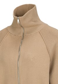 Beiger Sweatpullover mit Reißverschluss und hohem, geripptem Kragen, mit einem dezenten Logo auf der Brust, aus weichem Stoff mit glatter Textur gefertigt.