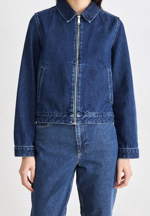 Femme portant une veste en denim bleu moyen avec une fermeture éclair devant et un jean assorti, debout devant un fond uni.