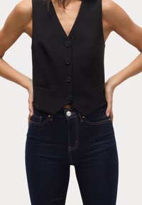 Chaleco negro con escote en V profundo, cinco botones y bolsillos laterales, combinado con unos vaqueros de tiro alto de color azul oscuro. Diseño minimalista y textura elegante.