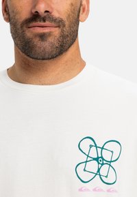 Hvid bomulds t-shirt med et teal abstrakt drone-design med et cirkulært mønster og pink accenter nær kanten.
