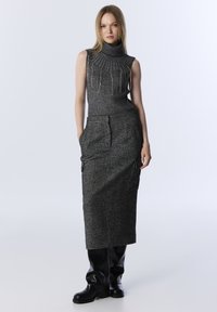 Vrouw in een mouwloze grijze coltrui met versieringen, lange grijze rok met zakken, en zwarte kniehoge laarzen.