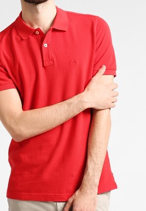 Homme portant un polo rouge à manches courtes tenant son haut du bras gauche avec sa main droite sur un fond uni.