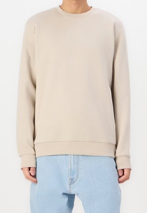 Personne portant un sweat-shirt beige à col rond avec poignets et ourlet côtelés, associé à un jean en denim bleu clair.