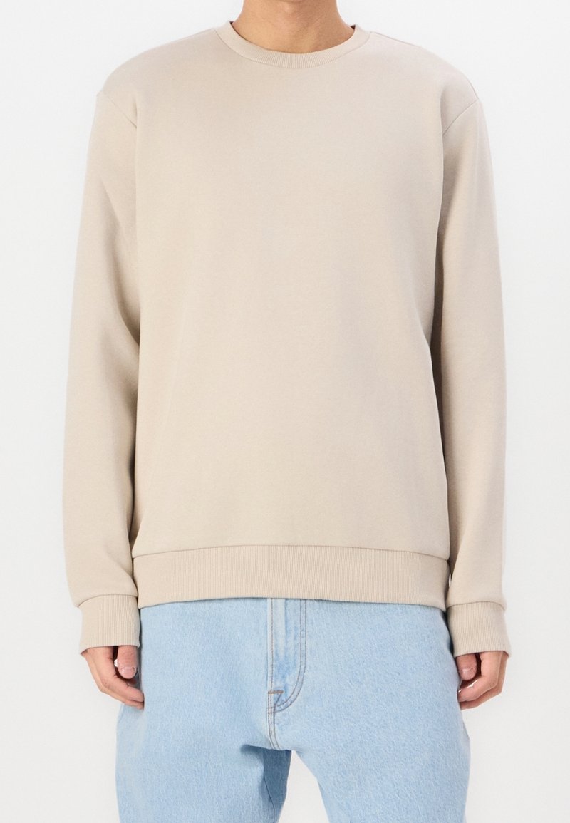 Personne portant un sweat-shirt beige à col rond avec poignets et ourlet côtelés, associé à un jean en denim bleu clair.