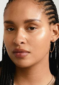 Primer plano de una cara con cabello trenzado, que lleva un aro de oro delgado y texturizado. Piel suave y clara con maquillaje natural y un color de labios neutro.