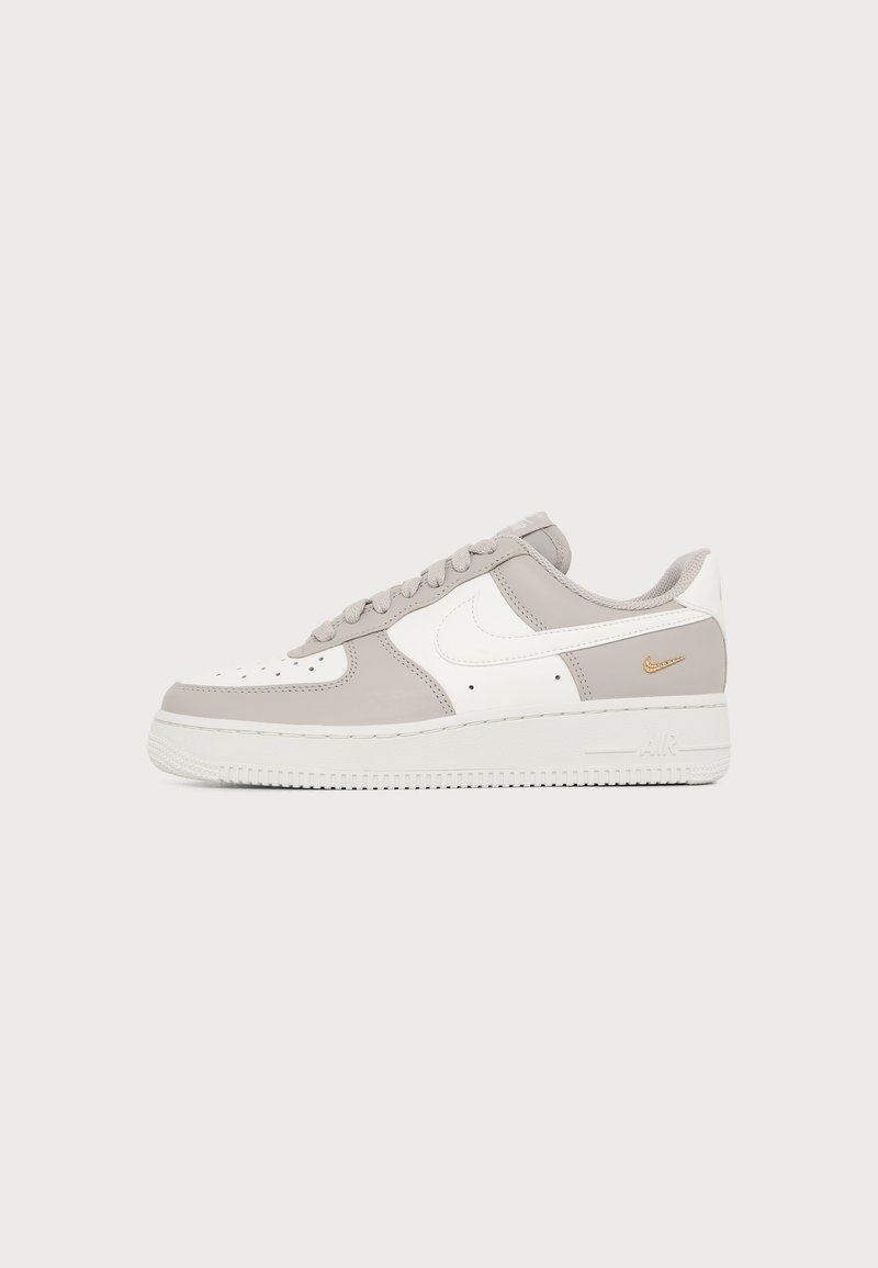 Χαμηλό αθλητικό παπούτσι Nike Air Force 1 σε λευκό και ανοιχτό μπεζ με λευκά κορδόνια και μικρό χρυσό λογότυπο Nike κοντά στη φτέρνα.