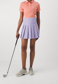 Kvinna med en rosa polo och en lila plisserad kjol som håller en golfklubba, stående mot en enkel vit bakgrund.