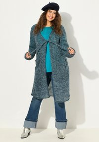 Lång tealfärgad cardigan med en mjuk, stickad textur, öppen design och framfickor. Modellen bär en blå topp och utsvängda blå jeans med silverstövlar.