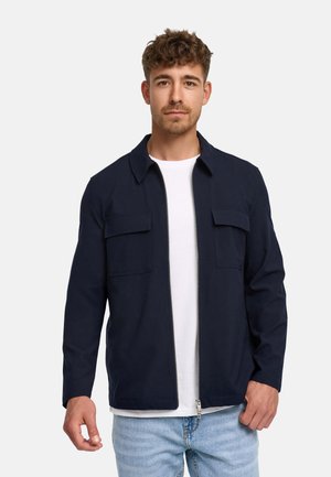 Mann mit kurzen lockigen Haaren, der eine marineblaue Jacke mit Reißverschluss über einem weißen T-Shirt und hellblauen Jeans vor einem einfarbigen Hintergrund trägt.