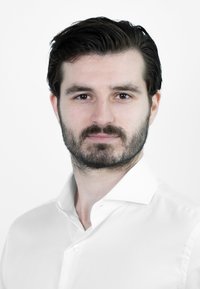 Portret van een man met donker haar en een baard, gekleed in een witte, knoopsluiting shirt met een zachte kraag. De achtergrond is simpel wit.