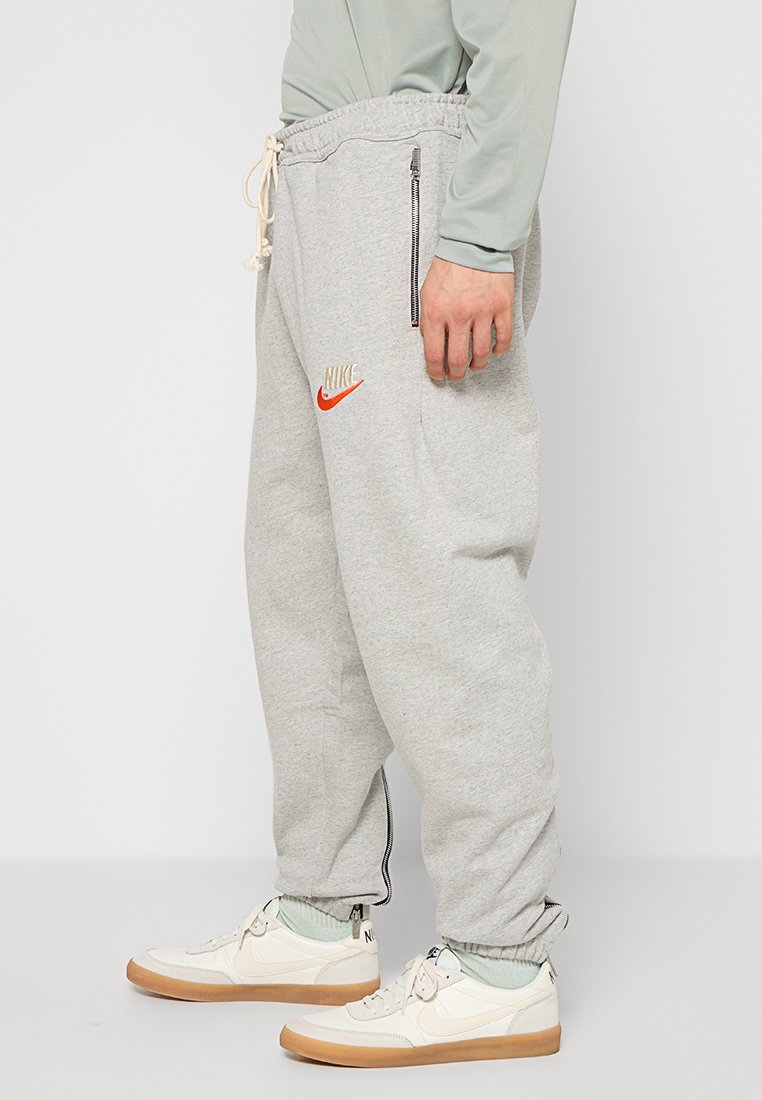 Jogging en gris avec taille ajustable, poches zippées sur les côtés et logo Nike en orange et crème. Porté avec des baskets blanches et des chaussettes pastel.