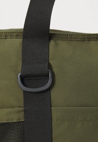 Sac en tissu vert olive avec une sangle en nylon noir. Comprend une boucle en plastique et un panneau en maille texturée. Les coutures mettent en valeur la structure.