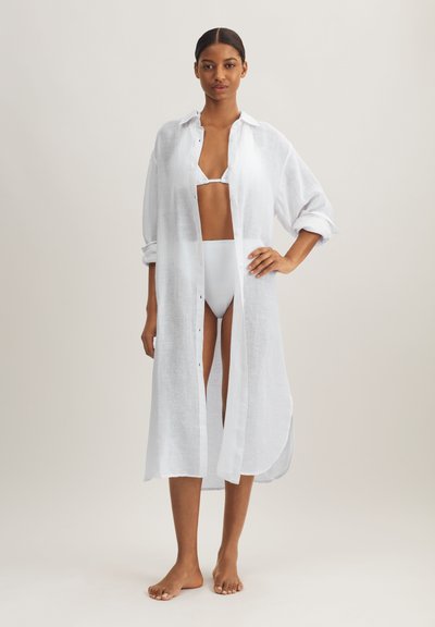 Λευκό, ελαφρύ λινό κάλυμμα με μακριά μανίκια και πλάγιες σχισμές, κλείσιμο με κουμπιά, φορεμένο πάνω από λευκό μπικίνι σετ που ταιριάζει.