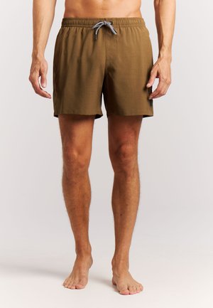 Man die bruine zwembroek draagt met een zwart-wit koord, staat op blote voeten tegen een effen achtergrond.