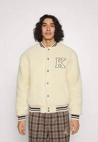 Cream-colored shearling jacket with black and white striped collar and cuffs, featuring a prominent "K" patch and snap button closure.