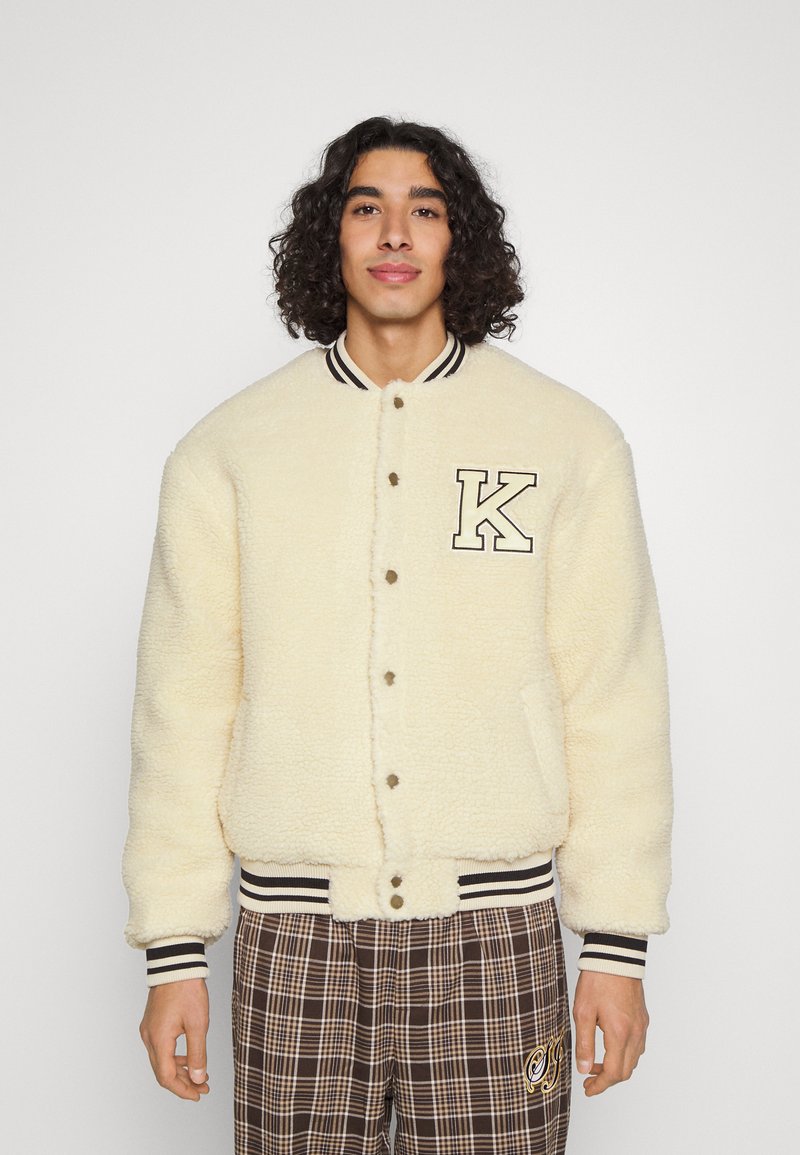 Cream-colored shearling jacket with black and white striped collar and cuffs, featuring a prominent "K" patch and snap button closure.
