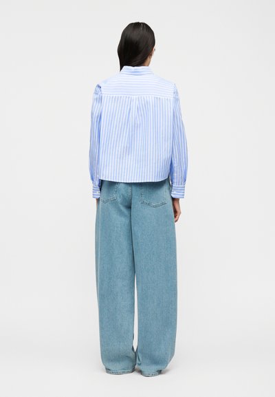 Γυναίκα που στέκεται με την πλάτη στην κάμερα, φορώντας μια μπλε και λευκή ριγέ μπλούζα με μακριά μανίκια και φαρδιά ανοιχτό μπλε τζιν παντελόνια.