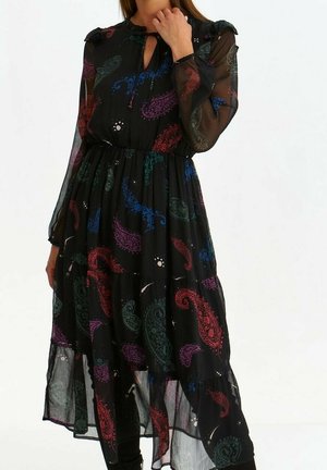 Kobieta ubrana w czarną, przezroczystą sukienkę na długim rękawie z wielokolorowymi wzorami paisley, z dekoltem typu keyhole i marszczoną talią.