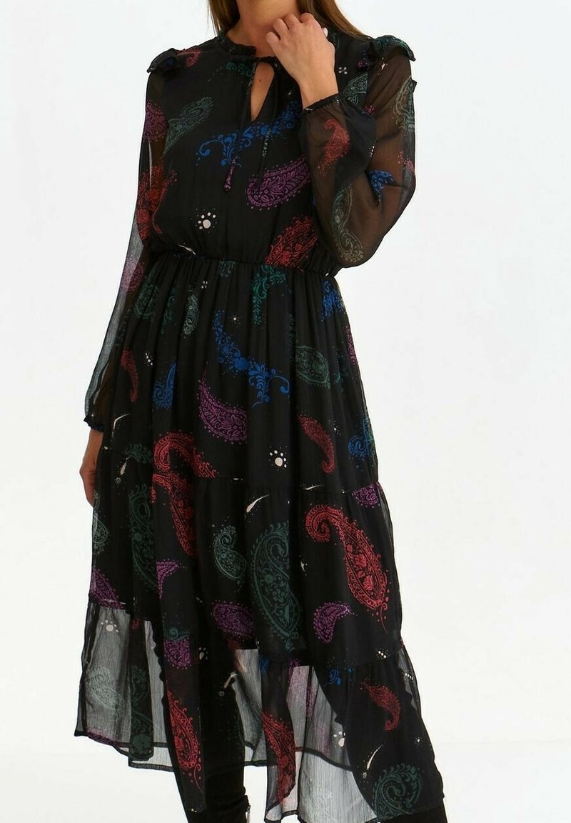 Femme portant une robe noire à manches longues en tissu transparent avec des motifs cachemire multicolores, dotée d'une encolure avec trou en forme de goutte et d'une taille froncée.