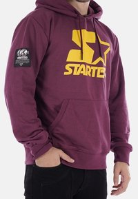 Sudadera color marrón con un gran logo amarillo de "STARTER", bolsillo tipo canguro en el frente y un parche negro en la manga izquierda. Material de mezcla de algodón.