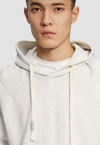 Sudadera gris claro con capucha ajustable, con una tela texturizada, mangas raglán y una pequeña etiqueta azul cerca del dobladillo.