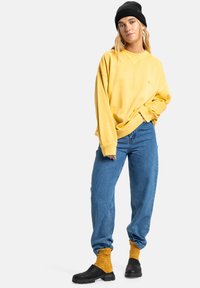 Geel sweatshirt met een relaxte pasvorm, blauwe denim jeans, zwarte beanie en zwarte instapschoenen. Geperforeerde gele sokken steken uit de jeans.