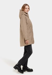 Beige, wasserdichter Parka mit Kapuze, ausgestattet mit zwei Seitentaschen, geradem Schnitt und glatter Textur, kombiniert mit schwarzen Stiefeletten.