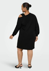 Čierne šaty so sweatshirt strihom, dlhými rukávmi, asymetrickým off-shoulder dizajnom, mäkkou textúrou a dĺžkou do kolien. Nosené s čiernymi sandálmi na podpätku.