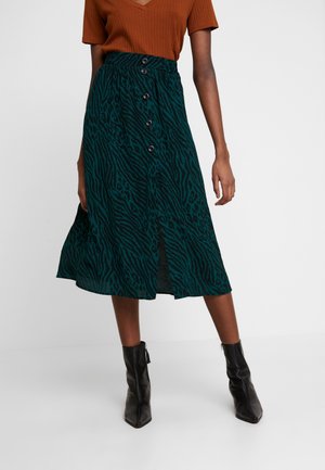 Femme portant une jupe midi imprimé animal vert et noir avec des boutons, un top marron à manches courtes et des bottines noires, debout sur un sol blanc.