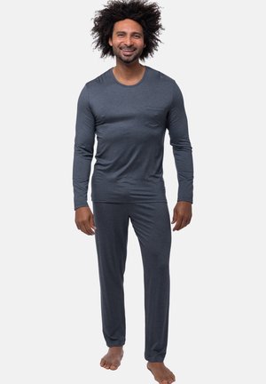 Man met krullend haar, gekleed in een donkergrijze top met lange mouwen en bijpassende broek, staat blootsvoets tegen een effen achtergrond.