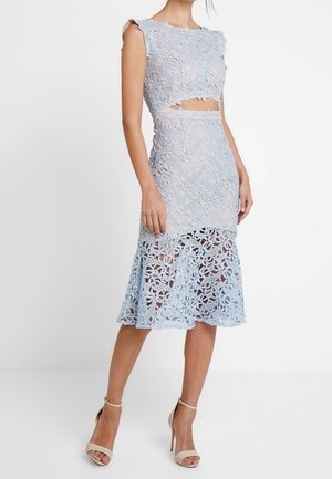 Kobieta ubrana w bezrękawową, jasnoniebieską koronkową sukienkę dwuczęściową z wycięciem na brzuchu i przejrzystą spódnicą z kwiatowej koronki, w połączeniu z beżowymi sandałami na wysokim obcasie.