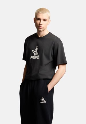 Młoda osoba z krótkimi blond włosami nosi czarną koszulkę i spodnie z białym logo Prosto, stoi z rękami w kieszeniach na jasnym tle.