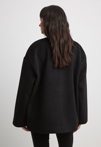 Czarna, oversize'owa kurtka z polaru z zaokrąglonym dołem, szerokimi rękawami i miękką teksturą, noszona na czarnych spodniach. Widok z tyłu nie pokazuje zapięć.