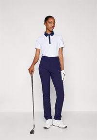 Bílý polo tričko s námořnickým límcem, spárované s námořnickými kalhotami. Model drží golfovou hůl a má na sobě bílé sportovní boty a rukavici.