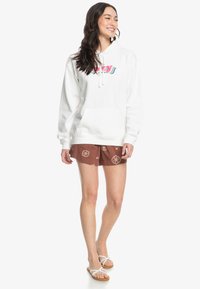 Witte hoodie met logo, voorzak en koordjes, gecombineerd met bruine gemustreerde shorts en witte sandalen. Gemaakt van een gemengde katoen met textuur.