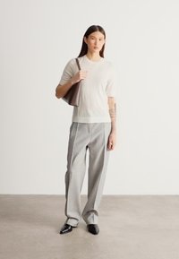 Kremowa dzianinowa bluzka z krótkim rękawem w połączeniu z szarymi spodniami w kratę. Modelka niesie brązową skórzaną torbę typu tote i ma na sobie czarne buty z noskiem w szpic.