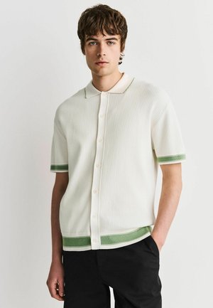 Junger Mann, der ein weißes kurzärmliges Hemd mit grüner Einfassung an Ärmeln und Saum trägt und mit einer Hand in der Tasche steht.